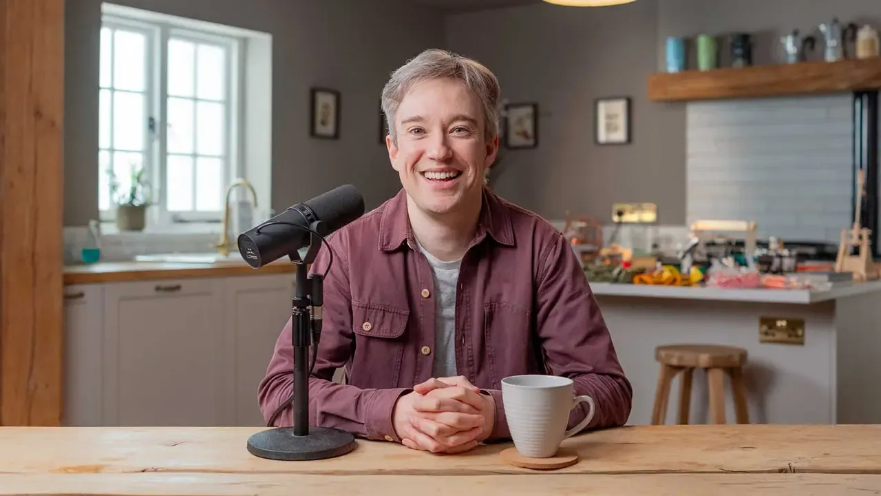 Tom Scott sitting at a desk with a microphone and a mug beside him.
